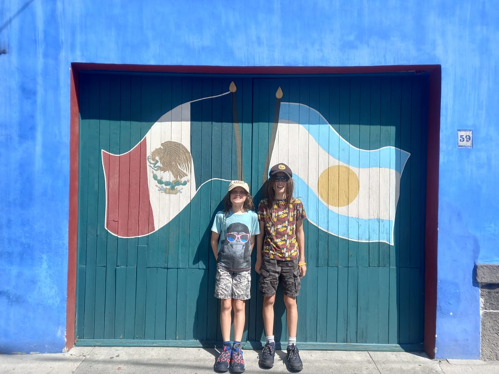 Two young boys stand in front of a flag mural in Mexico