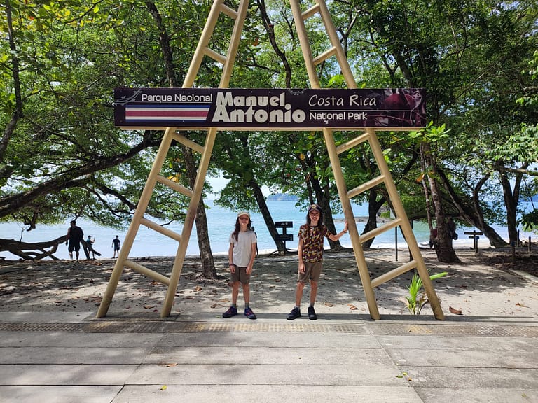 Two young boys stand in below a large sign saying Manuel Antonio National Park. Behind them are some trees and a beach